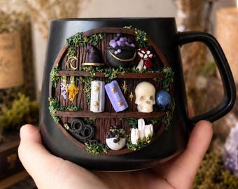 May include: A black ceramic mug featuring a circular, three-tiered shelf design decorated with miniature sculpted items including a witch hat, a cauldron with purple bubbles, books, a skull, a blue sphere, candles, and green moss accents.