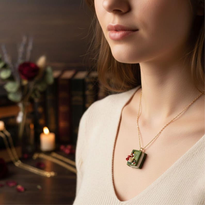 Tres collares con dijes en forma de libro miniatura de arcilla polimérica en colores crema, verde bosque y rojo cereza. Cada libro tiene flores en relieve (rosas y hojas) y una letra dorada en la portada. Las cadenas son de eslabones dorados y están dispuestas en diagonal sobre un fondo liso.