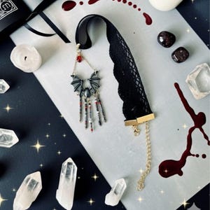 Choker hecho a mano con diseño de murciélago en tonos negro y gris, cuentas colgantes y lágrima roja, sobre fondo decorado con cuarzos, velas, piedras oscuras y detalles que imitan salpicaduras en rojo.