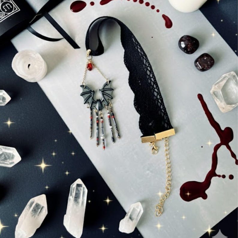 Choker hecho a mano con diseño de murciélago en tonos negro y gris, cuentas colgantes y lágrima roja, sobre fondo decorado con cuarzos, velas, piedras oscuras y detalles que imitan salpicaduras en rojo.