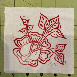 Pode incluir: Um desenho floral vermelho &eacute; impresso em um tecido branco. O desenho apresenta uma rosa com folhas e caules.