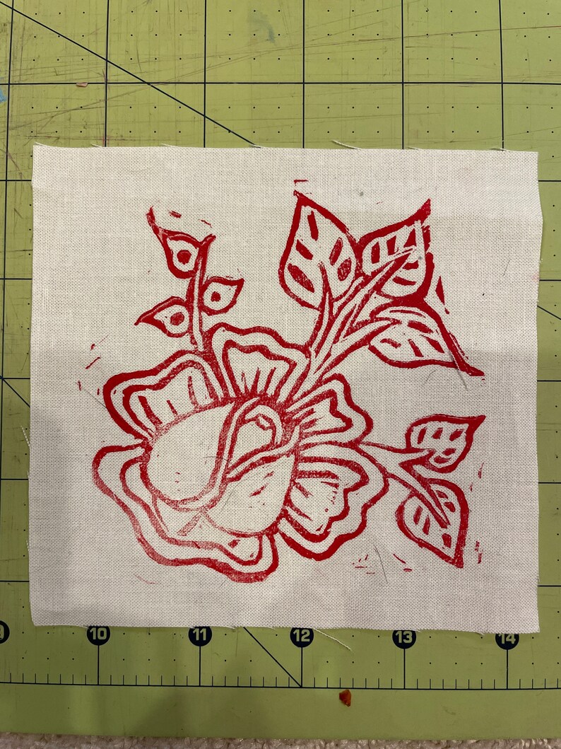Pode incluir: Um desenho floral vermelho &eacute; impresso em um tecido branco. O desenho apresenta uma rosa com folhas e caules.