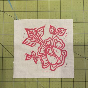 Pode incluir: Um desenho floral rosa impresso em um quadrado de tecido branco. O desenho apresenta uma rosa grande com folhas e caules.