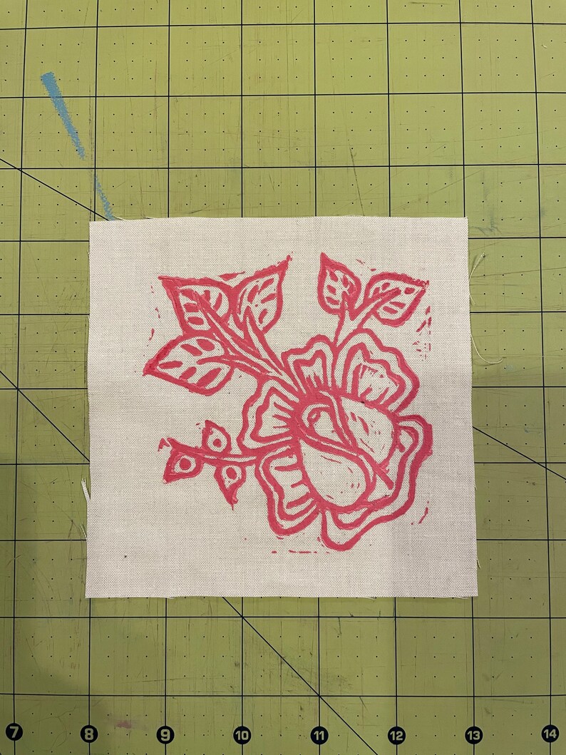 Pode incluir: Um desenho floral rosa impresso em um quadrado de tecido branco. O desenho apresenta uma rosa grande com folhas e caules.