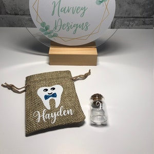 May include: A small hessian bag with a white tooth design and the name Hayden printed on it. A small glass bottle with a cork top is next to the bag. The bottle has a letter P written on it. A clear acrylic sign with a gold geometric design and the text Amber Harvey Designs is in the background.