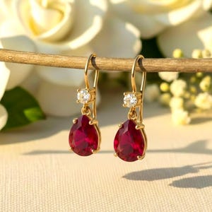 Peut inclure: Boucles d'oreilles pendantes dorées avec une pierre précieuse rouge foncé en forme de poire et une petite pierre transparente ronde. Les boucles d'oreilles sont suspendues à une branche en bois, avec des fleurs blanches en arrière-plan.