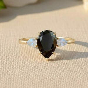 May include: A gold ring with a pear-shaped black gemstone as the centerpiece, flanked by two smaller, round, light-colored gemstones. The ring is set against a soft, neutral background with white flowers.
