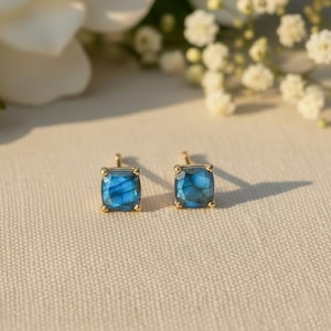 Peut inclure: Deux boucles d'oreilles à clous carrées en pierres bleues avec des montures dorées. Les boucles d'oreilles sont présentées sur une surface neutre, avec des fleurs blanches en arrière-plan. Ce sont des bijoux.