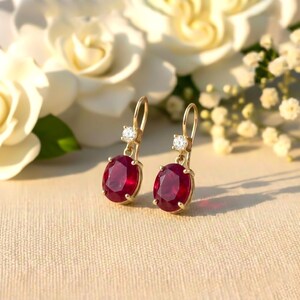 Puede incluir: Un par de pendientes colgantes dorados con piedras preciosas ovaladas de color rojo intenso y pequeñas piedras de acento claras y redondas. Los pendientes están colocados sobre un fondo suave y neutro con flores blancas.
