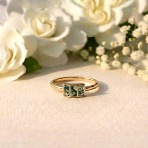 May include: A gold ring featuring three rectangular moss agate stones. The ring is set against a soft, white background with white flowers. A delicate piece of jewellery.