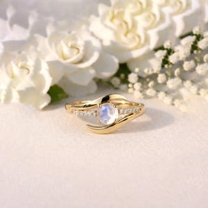 Puede incluir: Un anillo dorado con una piedra lunar central y pequeñas piedras de acento. El anillo tiene un diseño único y fluido. El fondo presenta flores blancas, creando una estética suave y romántica. El anillo se muestra sobre una superficie blanca.