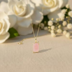 May include: A gold-colored necklace with a rectangular, light pink pendant. The pendant is set in a gold frame and hangs from a delicate gold chain. The background features white flowers.