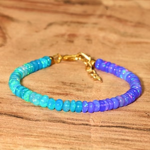May include: A beaded bracelet with a gradient of blue and purple opal stones. The bracelet has a gold-colored clasp and is displayed on a wooden surface. The beads transition from light blue to deep purple.
