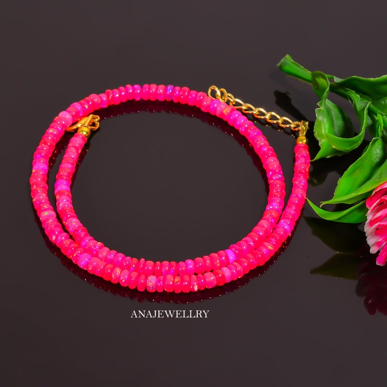 Puede incluir: Un collar de cuentas rosa vibrante con un cierre y cadena dorados. El collar est&aacute; compuesto por peque&ntilde;as cuentas redondas, creando un efecto en capas. El fondo es oscuro, resaltando el color brillante del collar. El texto "ANAJEWELLRY" es visible.