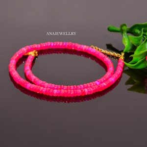Puede incluir: Un collar de cuentas rosa brillante con un cierre dorado. El collar est&aacute; compuesto por peque&ntilde;as cuentas redondas y se muestra sobre una superficie negra reflectante. El texto "ANAJEWELLRY" es visible en el fondo.