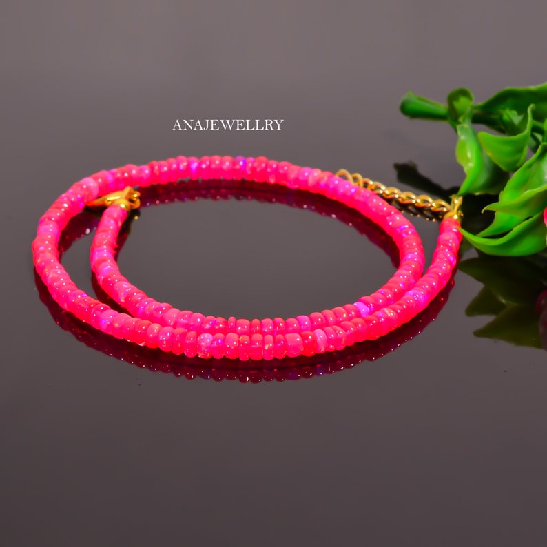 Puede incluir: Un collar de cuentas rosa brillante con un cierre dorado. El collar est&aacute; compuesto por peque&ntilde;as cuentas redondas y se muestra sobre una superficie negra reflectante. El texto "ANAJEWELLRY" es visible en el fondo.