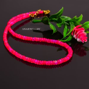 Puede incluir: Un collar de cuentas rosa brillante con un cierre dorado. El collar est&aacute; compuesto por peque&ntilde;as cuentas redondas, creando una hebra continua. El fondo es oscuro, resaltando el color brillante del collar. El texto "ANAJEWELLRY" es visible.