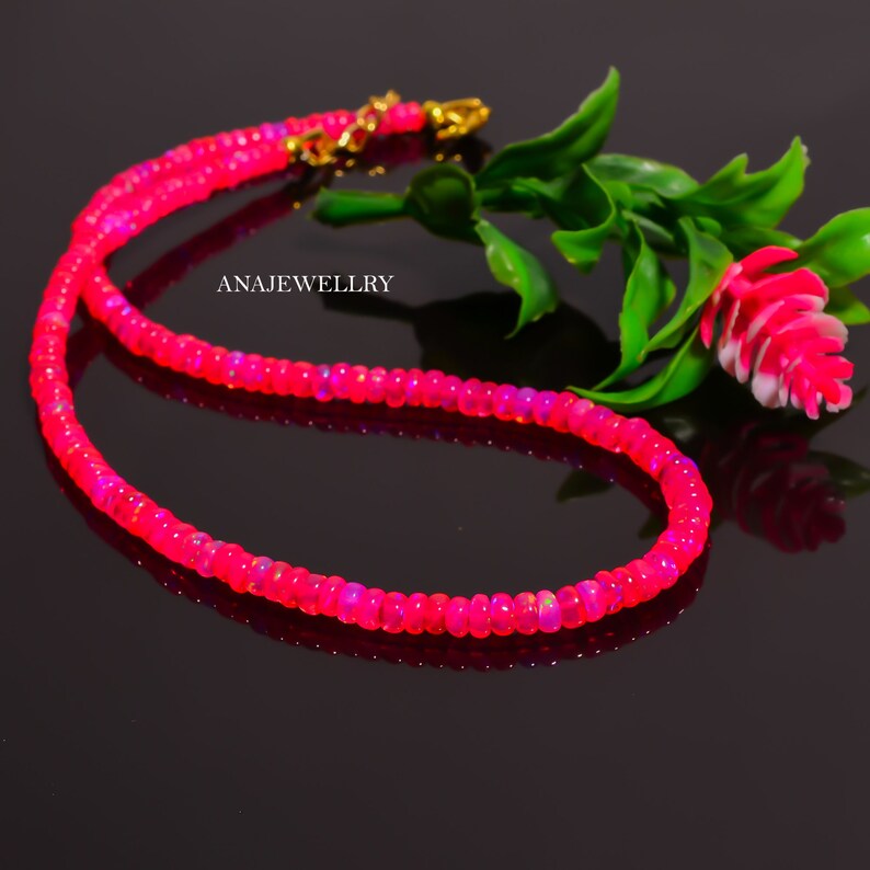 Puede incluir: Un collar de cuentas rosa brillante con un cierre dorado. El collar est&aacute; compuesto por peque&ntilde;as cuentas redondas, creando una hebra continua. El fondo es oscuro, resaltando el color brillante del collar. El texto "ANAJEWELLRY" es visible.