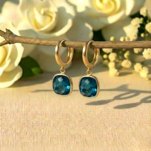 May include: A pair of gold hoop earrings with square, faceted, teal-colored gemstones. The earrings are suspended from a gold branch, with white flowers in the background. The earrings are a classic jewelry item.
