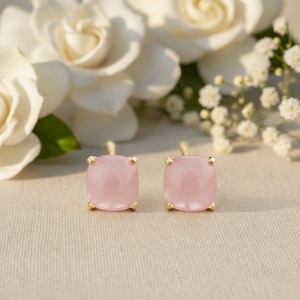 Puede incluir: Un par de pendientes de botón cuadrados de piedra preciosa rosa claro con garras doradas. Los pendientes están colocados sobre un fondo suave y neutro con flores blancas.