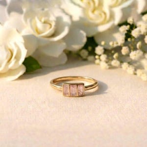 May include: A gold ring with three rectangular, light pink gemstones set in a row. The ring is displayed on a white surface, with white flowers in the background. The ring's band is thin and delicate.