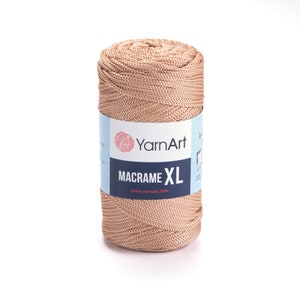 Puede incluir: Un carrete de cordón de macramé beige, etiquetado como "YarnArt Macrame XL" con el sitio web "www.yarnart.info" impreso en la etiqueta.
