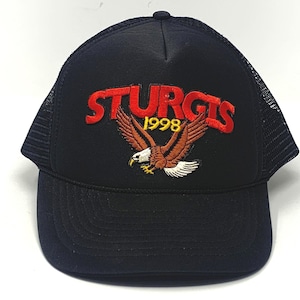 May include: Black trucker hat with a mesh back and a curved brim. The front features the word "STURGIS" in red, the year "1998" in gold, and an embroidered eagle with outstretched wings.