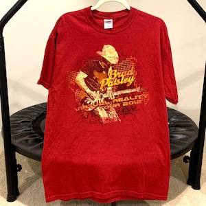 May include: Red t-shirt featuring a graphic of Brad Paisley playing a guitar, with the text "Virtual Reality Tour 2012." The shirt is hanging on a white hanger.