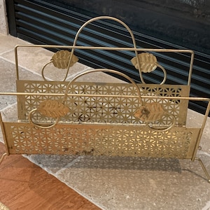 May include: A vintage gold-toned metal magazine rack. The rack features a rectangular design with a geometric pattern and leaf accents. It has a handle for carrying and is supported by slender legs. The rack is likely used for storing magazines or newspapers.