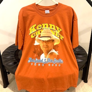 May include: Orange t-shirt featuring a graphic of Kenny Chesney wearing a cowboy hat. The text "Kenny Chesney" is arched across the top, with "The food & The radio TOUR 2006" below the image. The shirt is hanging on a hanger.
