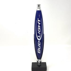 May include: A blue and silver Bud Light beer tap handle. The handle has a sleek, elongated design with the Bud Light logo in white. The base is a small, black square.