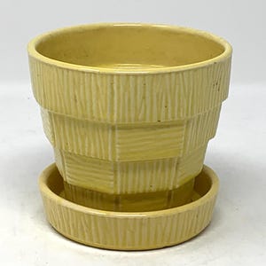 May include: A pale yellow ceramic planter with a matching saucer. The planter has a textured, stacked design. The saucer is round and has a similar textured pattern. This decorative pot is suitable for small plants.