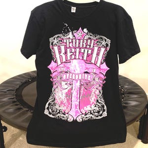 May include: Black t-shirt featuring a pink and white graphic design with the name "Toby Keith" prominently displayed. The design includes stars, ornate details, and the word "OVERDRIVE". The shirt is short-sleeved.