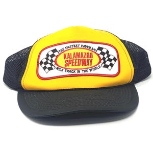 May include: Yellow and black trucker hat with the text "Kalamazoo Speedway" and checkered flags. The hat has a black mesh back and a curved black brim. The text on the hat reads "The Fastest Paved 3/8 Mile Track in the World."
