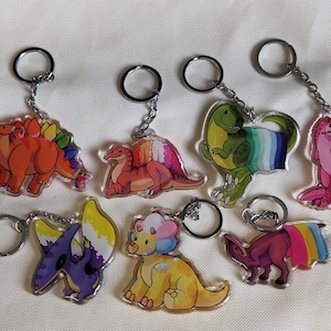 Może przedstawiać: Zestaw siedmiu kolorowych breloków z dinozaurami z tęczowymi flagami dumy. Każdy brelok przedstawia innego dinozaura z unikalnym wzorem i srebrnym kółkiem do kluczy.