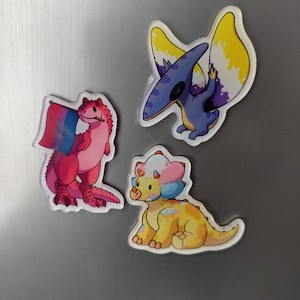 Può includere: Tre magneti di dinosauri colorati. Un dinosauro rosa con una bandiera blu, bianca e viola, un dinosauro giallo con accenti rosa e blu, e un pterodattilo blu e viola con ali gialle.