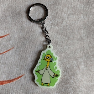 Pode incluir: Um chaveiro com um personagem de desenho animado verde e amarelo com uma corrente prateada. O personagem é uma figura amarela alta e fina com um pescoço comprido, olhos grandes e uma boca pequena. O personagem está vestindo uma túnica branca e está rodeado por uma aura verde e giratória.