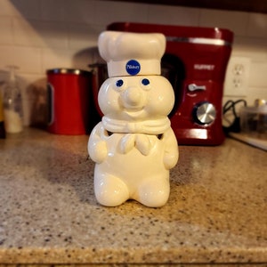 Peut inclure: Pot à biscuits Pillsbury Doughboy en céramique blanche. Il porte un chapeau de chef et un logo Pillsbury bleu. Le pot a un visage souriant et est posé sur un comptoir moucheté.