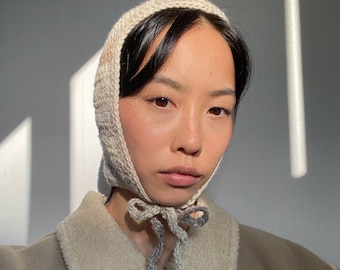 May include: A person wearing a beige knitted hooded hat with a long tie. The hat is tied at the neck and the person is wearing a beige fleece jacket.