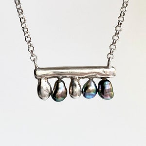 May include: A silver chain necklace featuring a horizontal silver bar pendant. Suspended from the bar are five small, teardrop-shaped charms, alternating between polished silver metal and dark, iridescent, multi-colored pearl-like beads.