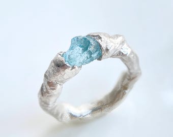 May include: A ring featuring a raw, light blue gemstone set within a textured, metallic silver band. The band has an uneven, organic surface finish that wraps around the finger, securely holding the central, jagged-edged blue stone in place.