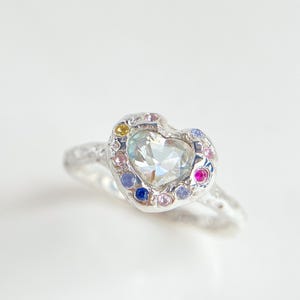 Può includere: Un anello d'argento con un cuore di pietra preziosa trasparente. Il cuore è circondato da un alone di piccole pietre preziose colorate. La fascia ha un design strutturato. L'anello è su sfondo bianco.