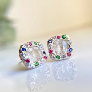 May include: A pair of silver earrings with a square-shaped, clear gemstone surrounded by a halo of smaller colorful gemstones. The gemstones are red, green, and blue.