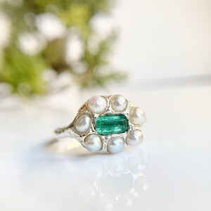 May include: A silver ring featuring a rectangular, vibrant green emerald gemstone surrounded by a halo of small, round pearls. The ring's band has a textured, organic design, and the setting is silver.