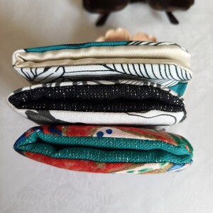 May include: A stack of three fabric pouches with different patterns. The top pouch is white with black lines and a teal border. The middle pouch is black with a white pattern and a teal border. The bottom pouch is red with gold and teal patterns.