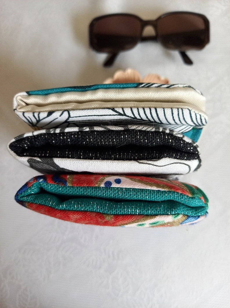 May include: A stack of three fabric pouches with different patterns. The top pouch is white with black lines and a teal border. The middle pouch is black with a white pattern and a teal border. The bottom pouch is red with gold and teal patterns.