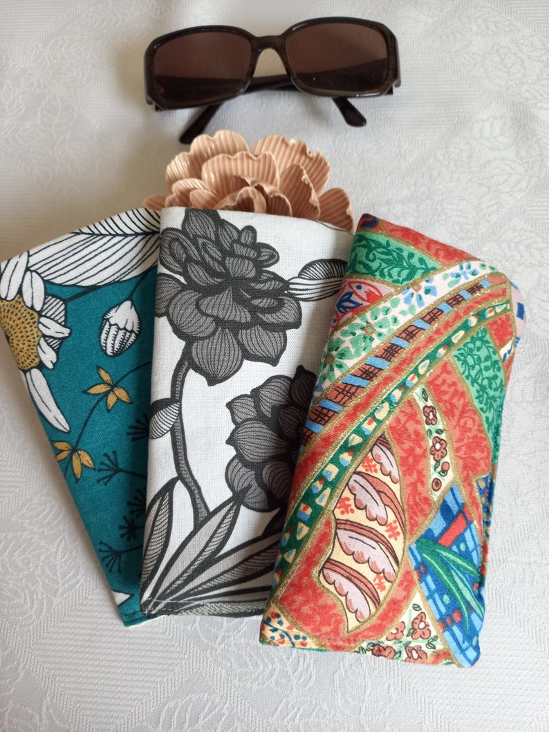 May include: Three fabric eyeglass cases with different patterns. The case on the left has a teal background with white and gold floral designs. The middle case has a white background with a black floral design. The case on the right has a red background with a colorful paisley design.