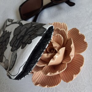 May include: A black and white floral patterned glasses case with a black zipper and a pink ceramic rose on a white surface.