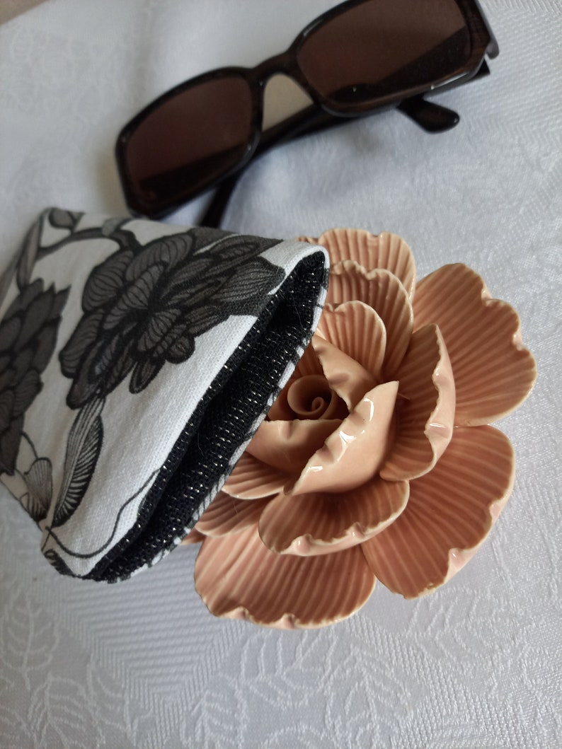 May include: A black and white floral patterned glasses case with a black zipper and a pink ceramic rose on a white surface.