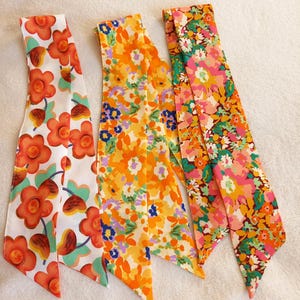 Puede incluir: Tres lazos para el pelo de tela con estampado floral en naranja, amarillo y rosa. Los lazos están atados en forma de lazo.